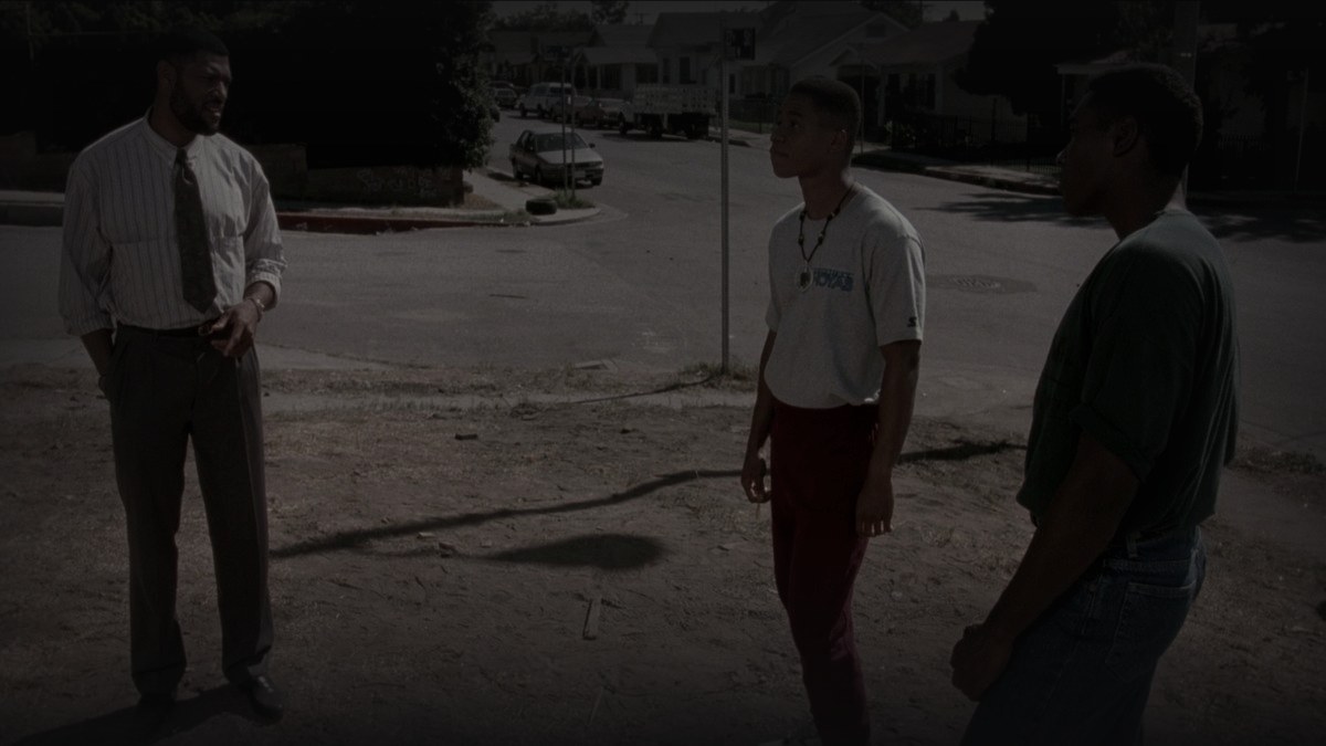 Still from Boyz n the Hood (1991) — 16:9 still from letterboxd (1200x675 crop variant), baked through scripts/bake_dark_photo.py at default factor 0.45. source luma 91.8, baked mean brightness 32.5 (in canon daylight range). scene: furious styles (laurence fishburne) talking to tre (cuba gooding jr.) and ricky (morris chestnut) on a south central street corner. cross-checked against tmdb backdrops; the letterboxd still is a genuine film frame, not promotional photography.